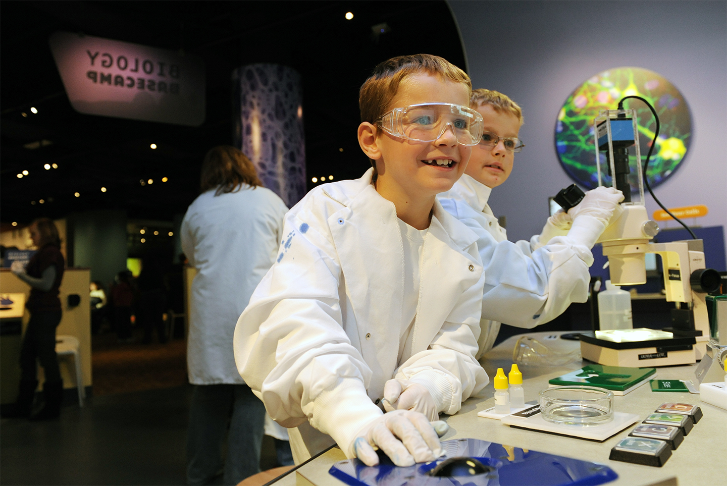 Two young boys wearing white lab coats, gloves, and safety goggles working with scientific equipment that is part of a museum exhibit