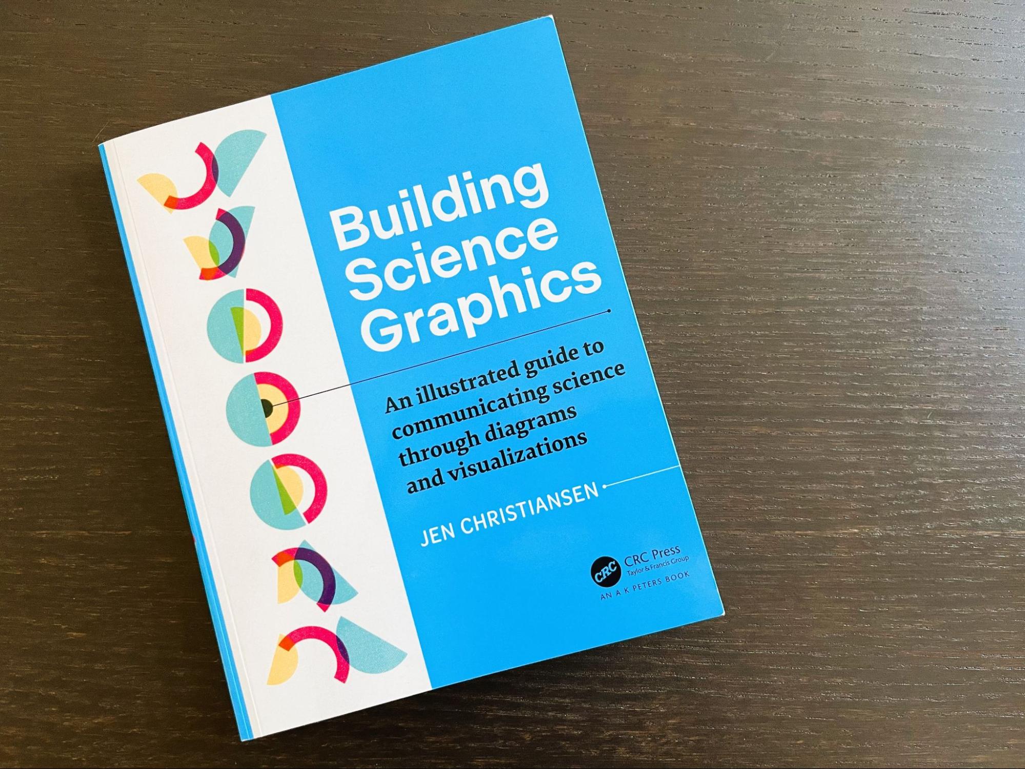 The Building Science Graphics book on a dark brown wood table. The book has a cover that is two-thirds blue and one-third white. On the white section of the cover, a series of overlapping half circles in yellow, pink, and blue are shown in varying degrees of rotation in a vertical arrangement.
