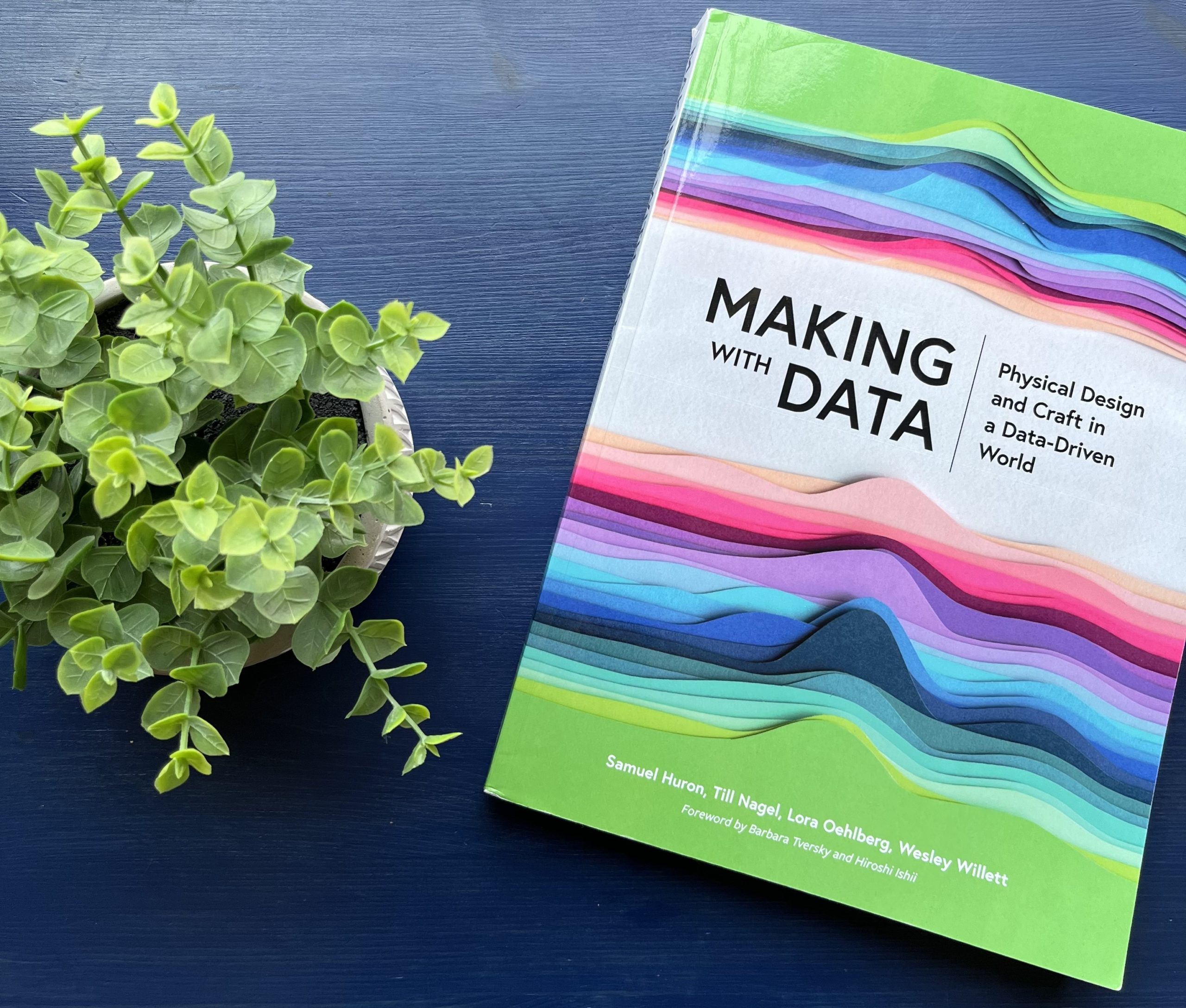 A colorful book sits on a blue table next to a small plant. The book’s cover shows a visualization made of many different colors of paper showing distribution curves. About a third of the way down the cover is the title “Making with Data Physical Design and Craft in a Data-Driven World.”