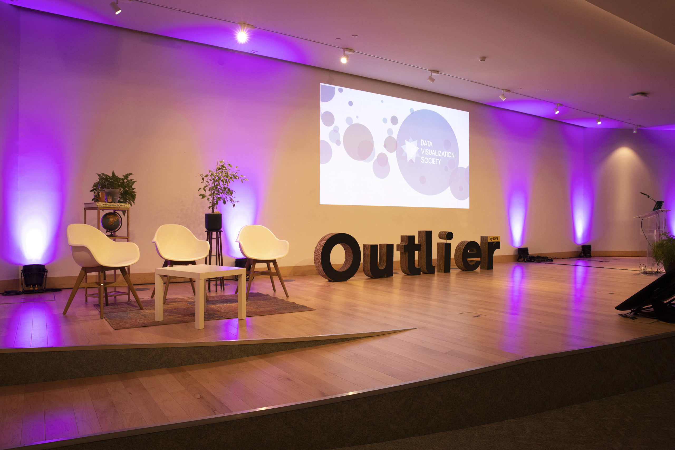 A photo of the Outlier presentation stage in Porto, with a projector screen three chairs, a podium and an physical "Outlier" letter blocks set on the floor.