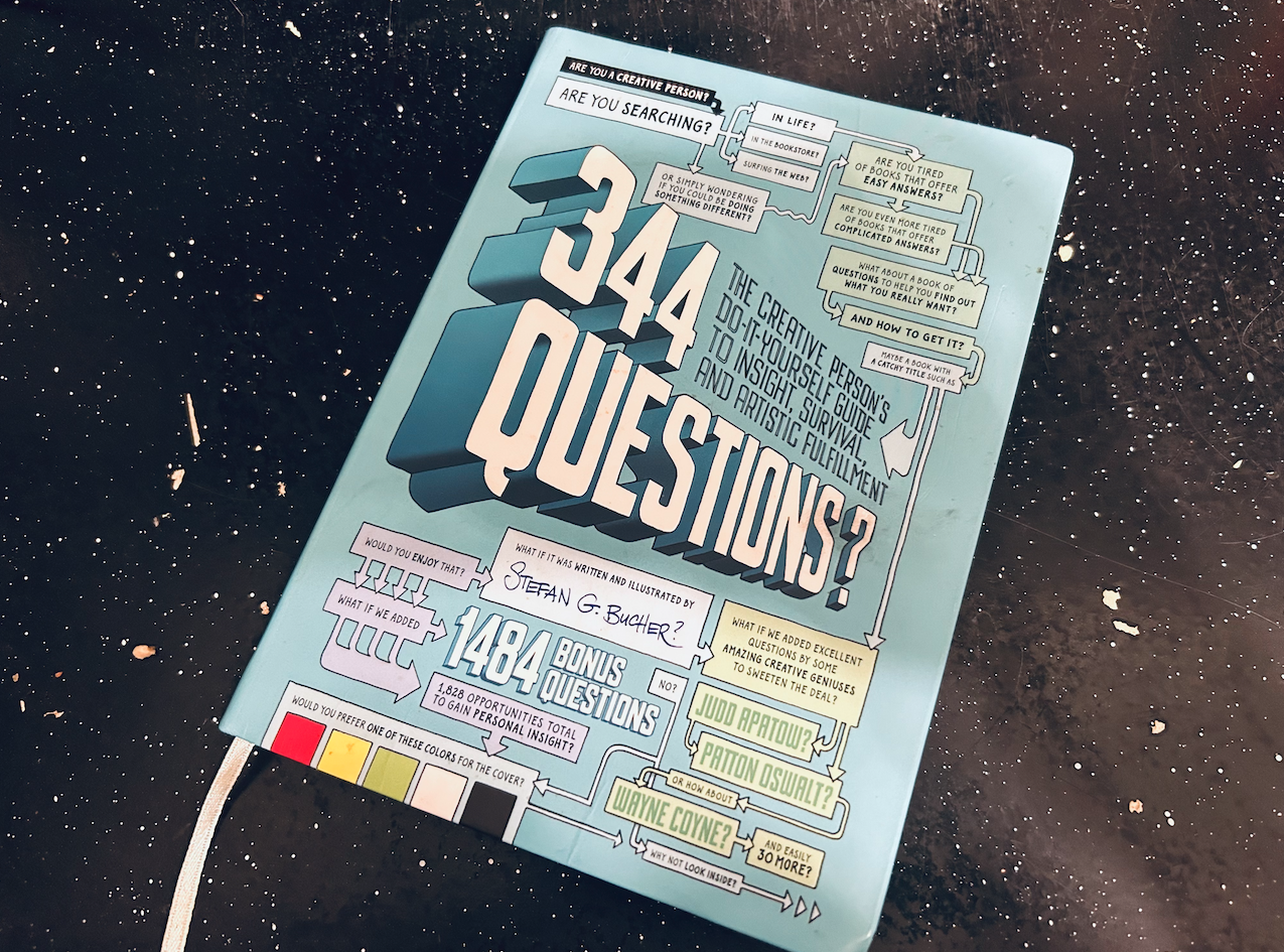 A baby blue book cover on a black, white-spattered tabletop. The book reads 344 Questions? The Creative Person's Do-It-Yourself Guide to Insight, Survival, and Artistic Fulfillment.