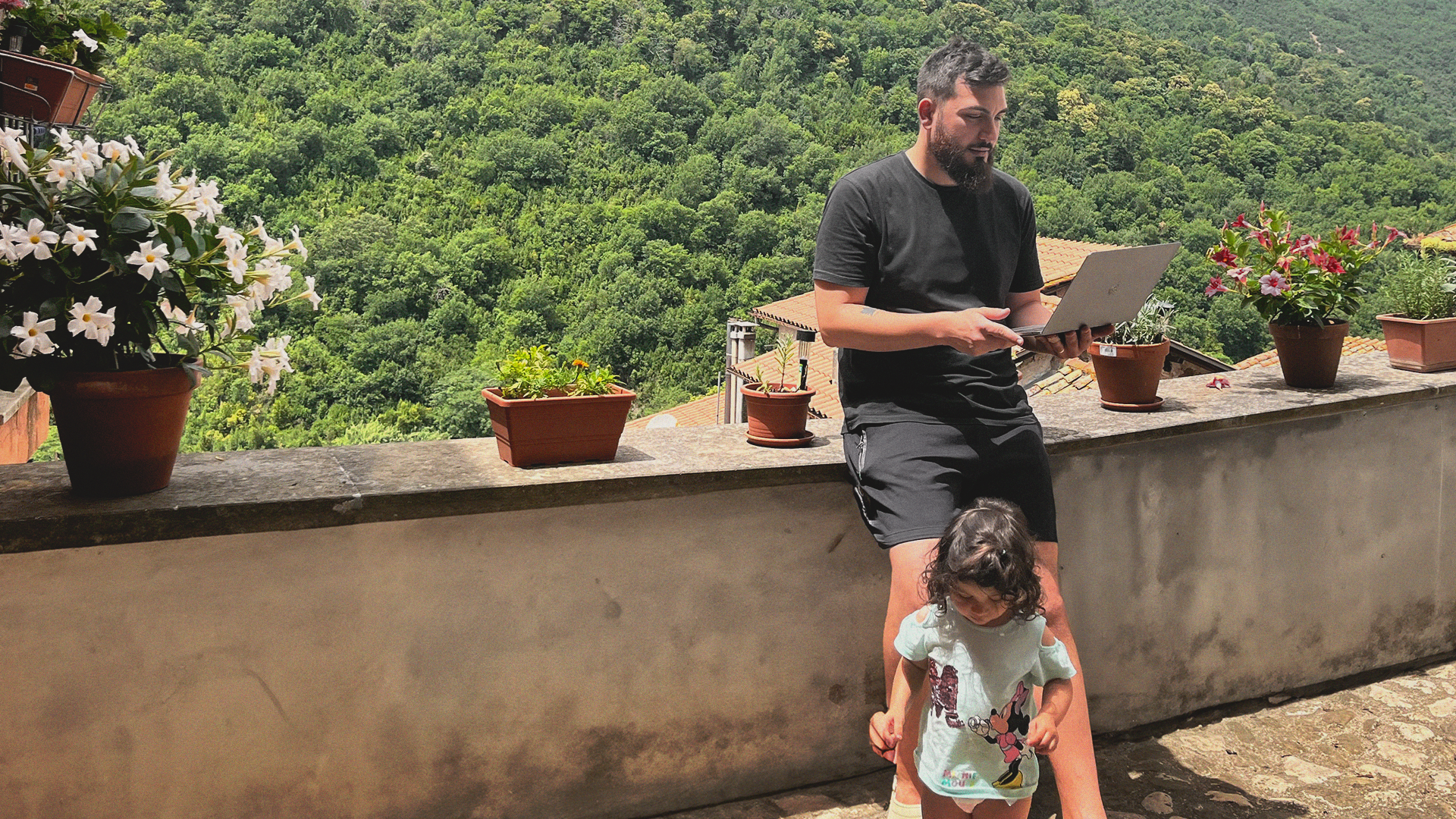 A man in the countryside holds a laptop while standing, his daughter wrapped around his legs.