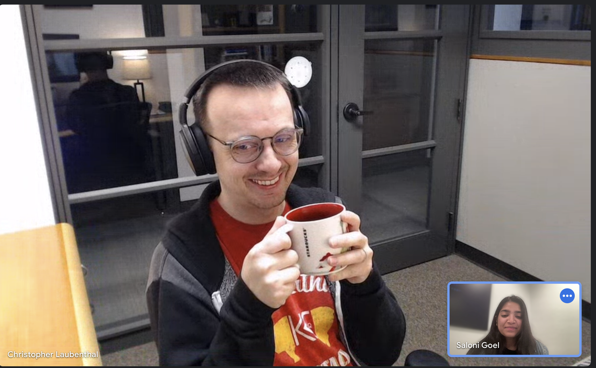 This image shows a virtual mentorship meeting between two individuals. The main focus is on a man wearing headphones and glasses, smiling warmly while holding a mug with both hands. In the bottom right corner, a smaller video frame shows a woman, his mentee, with her eyes closed and a slight smile on her face, engaged in the conversation. The atmosphere seems relaxed and friendly, suggesting a positive and comfortable mentoring relationship.