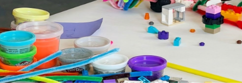 A workshop table covered in craft supplies: open tubs of Play-Doh, loose pipe cleaners, scattered Lego bricks, and a pencil, with colorful models built by participants in the background.